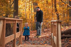 A toddler looks at a wooden bridge in a autumn forest as her father looks on.