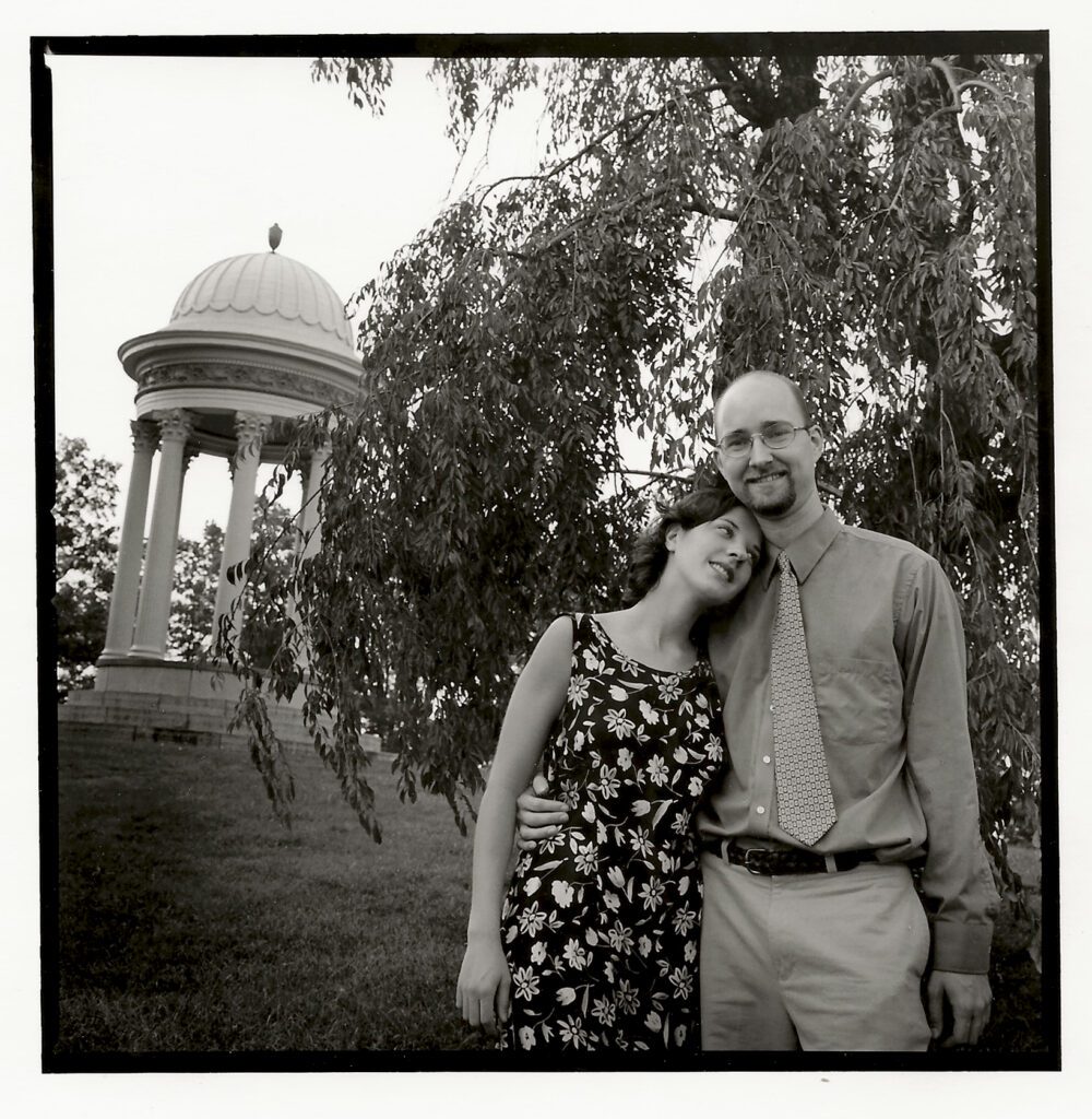 A young woman rests her head on the shoulder of a young man in front of a columned rotunda at a park.