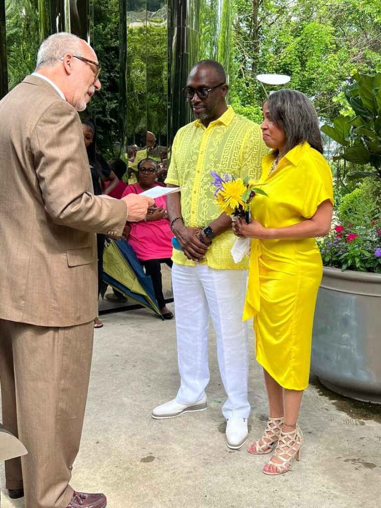 A man and woman dressed in yellow standing in front of a man in a beige suit, reading from a book.