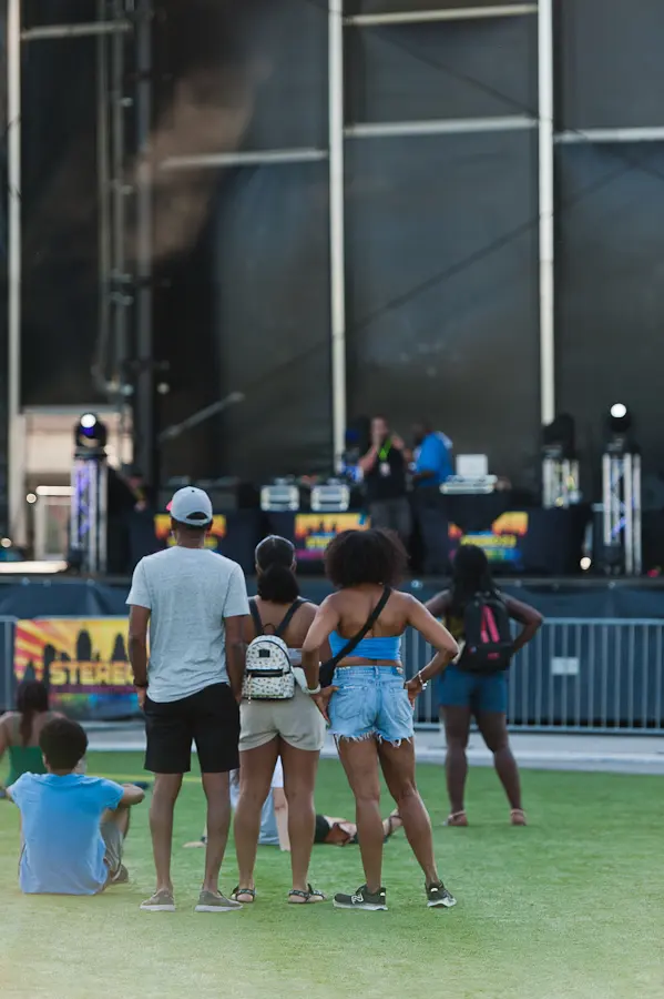 People are standing in green grass. They are facing a stage. There are people on the stage with microphones who are playing music.