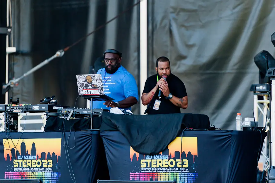 Two men standing on a stage. They are standing in front of DJ equipment and are talking on microphones.