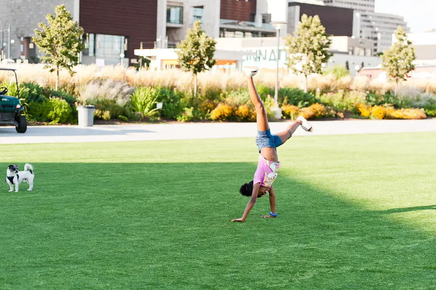 A child wearing shorts and a pink shirt, doing a handstand in the middle of a green lawn in a park. There are city buildings behind her.