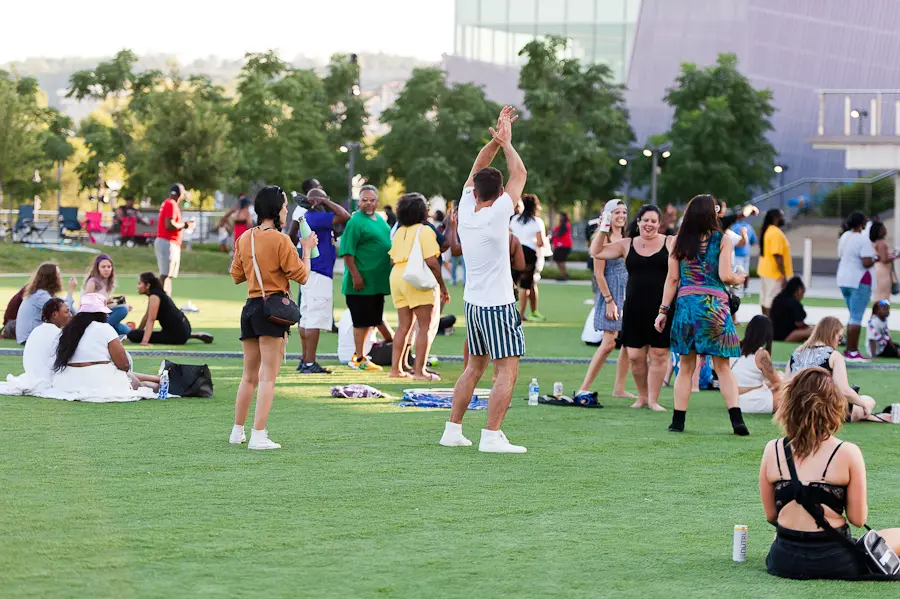 People gathered on a green lawn in a park outdoors. Some are sitting on blankets and other are dancing.