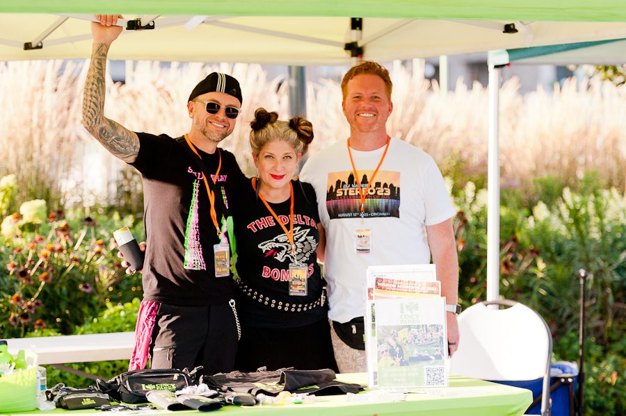 Three people posing for a picture. A man with a hat and tattoos is raising his arm to wave hello. He is standing next to a woman in a black shirt and next to her is a man with a white shirt. They are smiling.