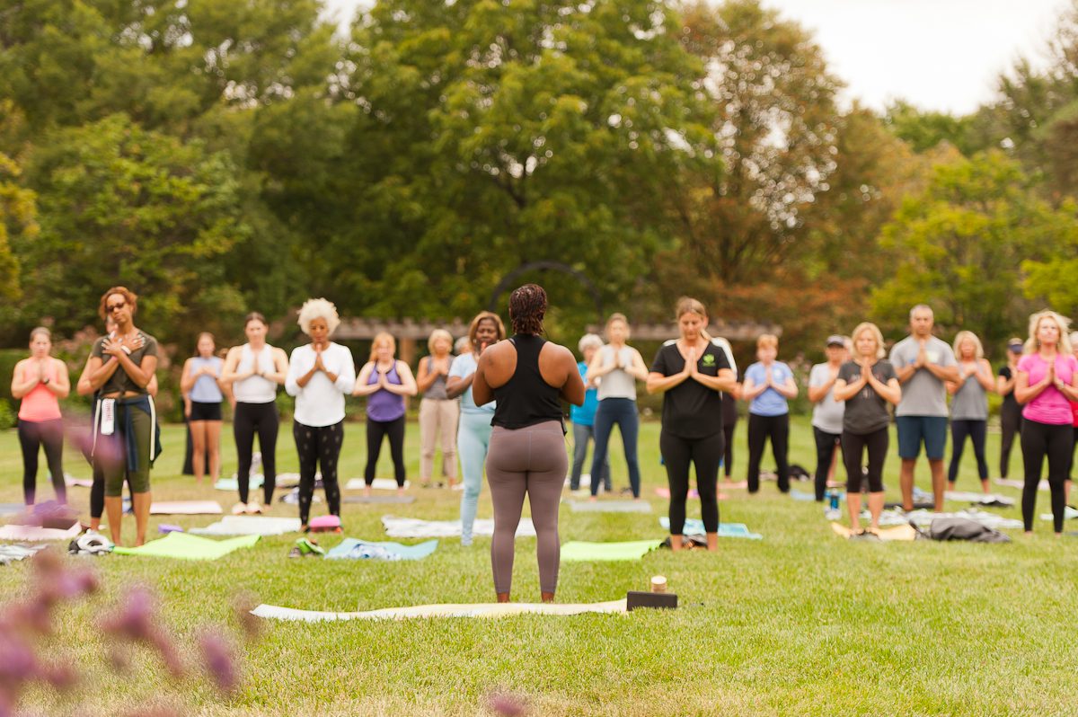 A large group of people and an instructor taking a yoga class on a large lawn in an outdoor park setting.