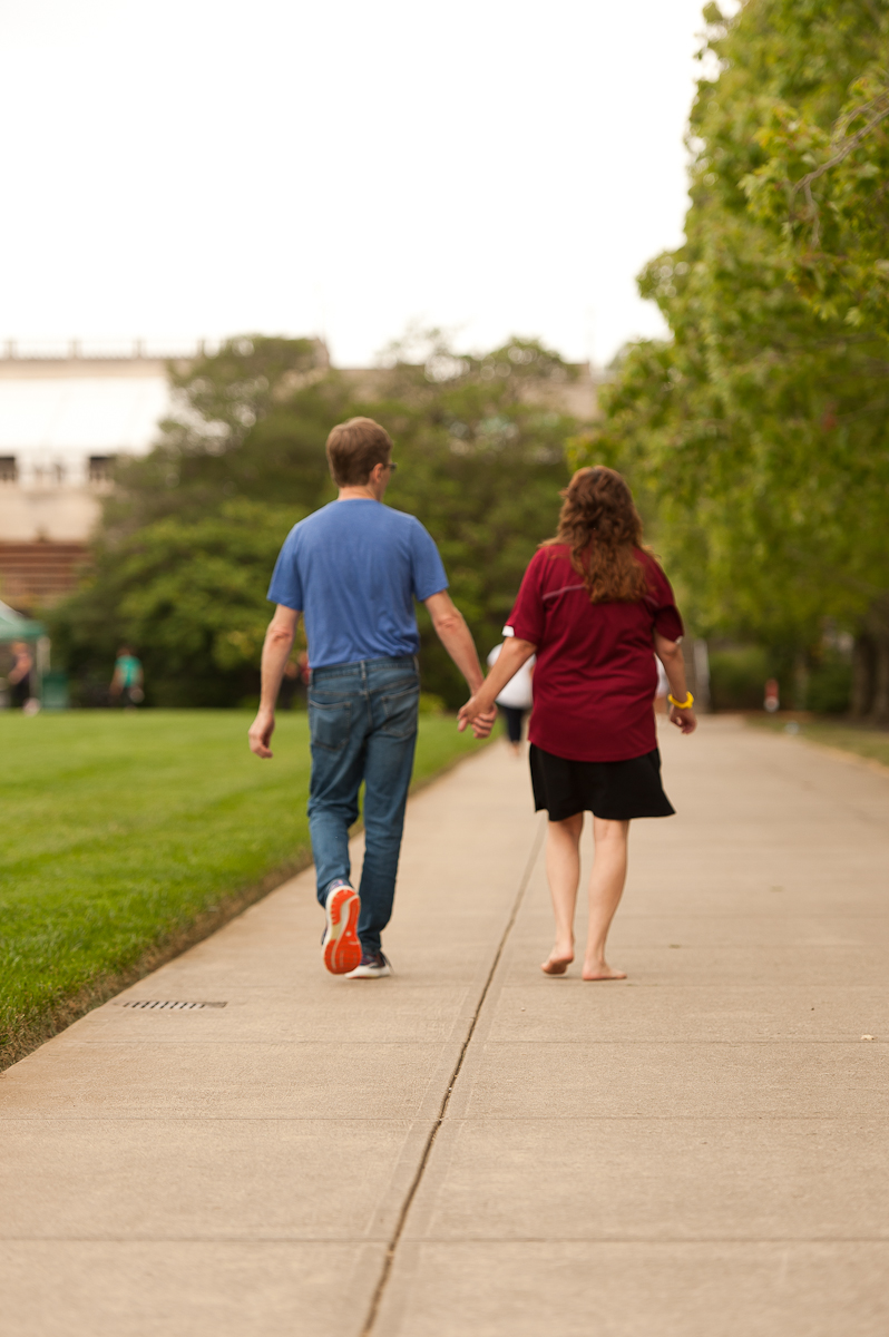 A man in a blue shirt and a woman in a red shirt are walking away from the camera, holding hands. They are walking along a sidewalk next to a green lawn and towards a group of trees.