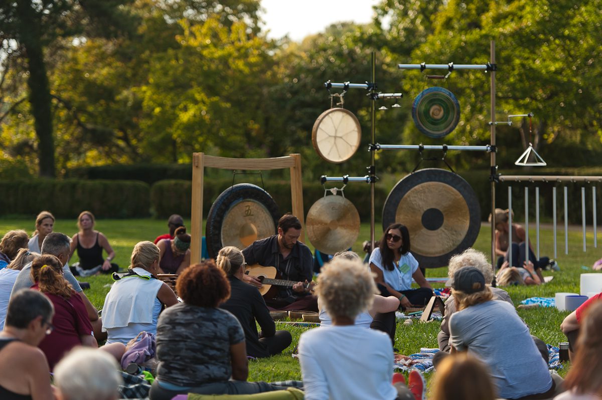 People sitting in front of a collection of gongs, chimes and other musical instruments in an outdoor park setting.