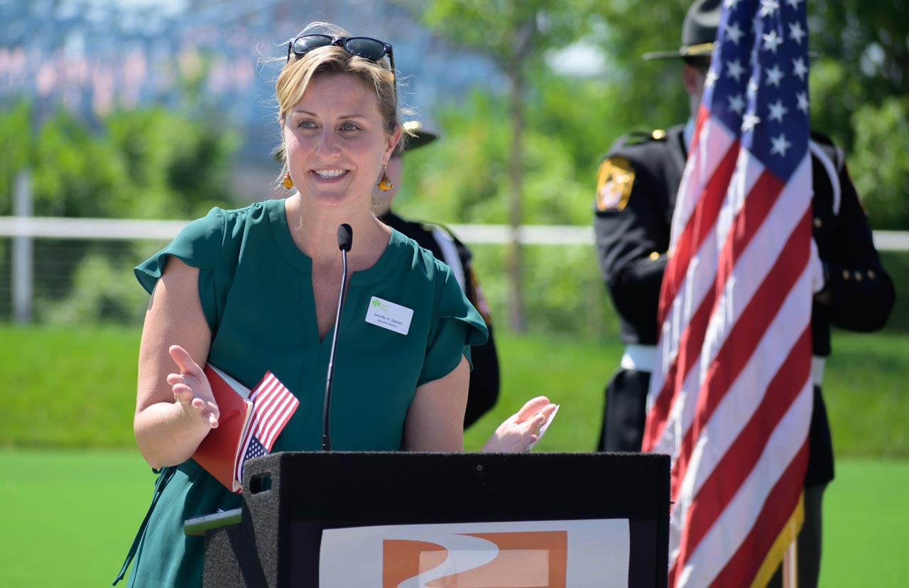 Jennifer Spieser is standing in a park. There are trees and green grass in the background. She is in front of a podium next to an American flag and is speaking into a microphone