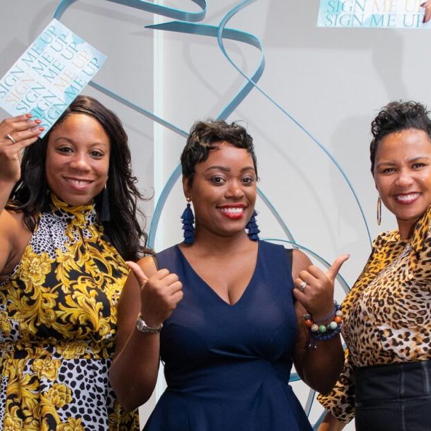 Three smiling women holding "sign me up" cards at a Women's Committee event